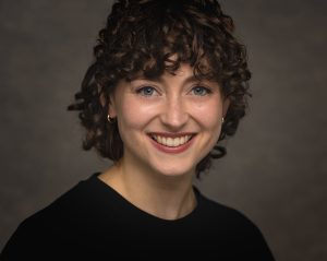 Profile image of a white female in her 30's. She has dark brown, curly, short hair, blue eyes and is wearing a black round-necked top. The background to the image is plain and brown.
