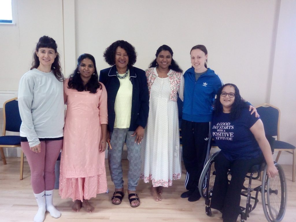 An image of 6 women of different ages and ethnicity standing together in a line, with the female to the right of the image in her wheelchair. They all have their arms linked behind and around each other and are smiling. Surroundings: a bright studio with pale wooden flooring with royal blue chair along the back wall
