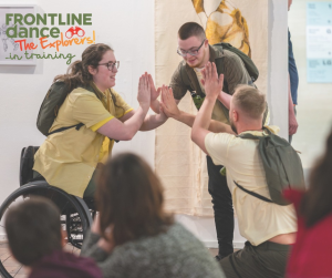 The Three Explorers are facing each other in a circle with the palms of their hands connected with each other. Beth is in her wheelchair, Connor is kneeling down, and Ben is standing up but leaning forward. They are all wearing different shades of yellow and khaki-green costumes and a backpack. The audience can be seen in the foreground.