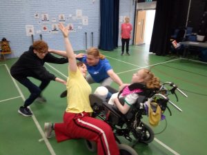 Breakthrou'Family session in a sports hall with green flooring and light blue walls. Dancers are all connected in a circle by their outstretched arms. One female in her wheelchair, two males crouched low and another female with one arm stretched to the ceiling. The facilitator is stood to the back of the room, watching.