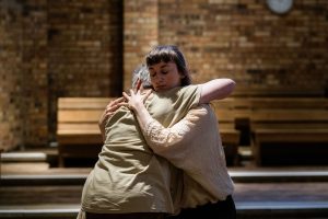 Image of two females, one with short grey hair and the other with long black hair with a short fringe, both wearing different shades of beige. They are holding each other in an embrace where one of their faces can be seen, and her eyes are closed, the other facing backwards, both are resting their heads on each other's shoulders. The image behind them is blurred out, but you can see a brown brick wall, a light wooden bench and a dark wooden flooring.
