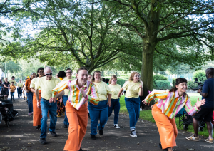 In a sunny park, the FRONTLINEdance are parading their routine amongst the trees and audience. They are all wearing bright yellow, orange and blue costumes, with big smiles on their faces.