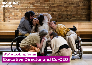 Five female dancers, two in their wheelchairs, are grouped together in a position where all are holding a different part of the central persons body. The image is predominantly in shades of beige and dull yellows and is set in a chapel, with a brick wall behind the dancers.