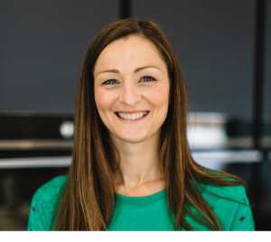 A profile image of Kirsten. She has long, brown, straight hair and wearing an emerald green, long-sleeved top with a rounded neckline. She stood behind a dark, blurred out background and has a beaming smile