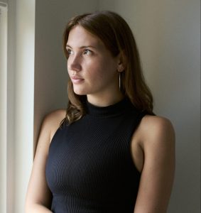 A profile image of Melanie. She has long, brown, wavy hair and is standing, gazing out of a window to her right. She is wearing a black, sleeveless, ribbed top and long silver earrings.