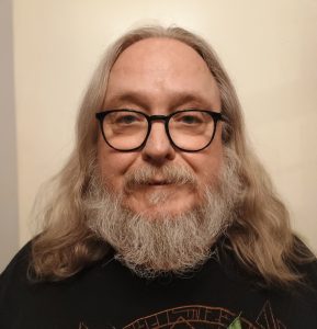 A profile image of Colin, who has long, wavy grey hair, a moustache & beard. He is wearing black rimmed glasses and a black jumper. He is looking directly forwards with a slight smile.