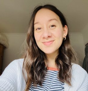 A profile photo of Jasmine, she has long, brown, wavy hair, and is wearing a blue & white striped top with a red rimmed neckline. Over the top, a white knitted cardigan. She is looking directly at the camera with a gentle smile.