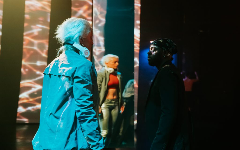 A white female with white short hair, wearing a turquoise jacket and headphones placed around the back of her neck, is looking towards a black male, wearing a black bandana and a black top. Both are standing in front of a mirrored background, and their reflections can be seen.