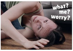 Kayleigh is lying on a wooden floor in front of a pale grey sofa. She is on her side, looking upwards with a worried expression. One arm is lay across her body, and the heel of her hand is resting adjacent to her forehead. Kayleigh is a white female with dark brown hair and is wearing a green vest top and blue jeans.