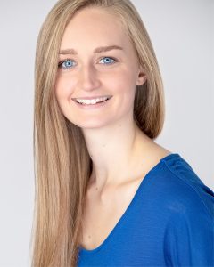 Profile image of Shannon: She has long, blonde, straight hair, falling over her right shoulder. She has blue eyes and is wearing a blue V-neck top. Background is very pale grey.