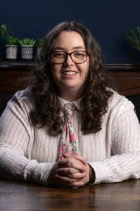 Profile photo of Sophie - she is sat at a desk, hand clasped in front of her. She has long brown curly hair, is wearing glasses and smiling. She has a light pink, striped, long-sleeved shirt on and a flowery pink & green tie. Behind Sophie is a dark brown wooden cabinet with three plant pots placed on the left side and one partially seen to the right.