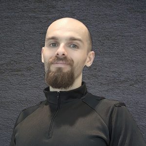 Profile image of Stanislav. He is standing in front of a black background and is wearing a black high-necked, zip-up top. Stan has a closely shaved head with a long goatee beard and is smiling.