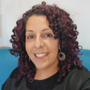 A profile image of Dana. She has long, dark brown, curly hair with red highilights. Dana is wearing a black top and silver hoop earings, she has brown eyes and is smiling. In the background of the image, there is a white wall and the top of a turquoise seat that Dana is at on.