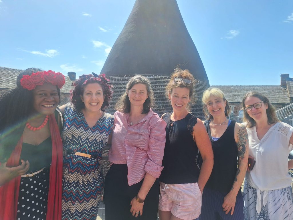 Six Parent Carer Artists all stood next to each other, in front of the bottleneck kiln at the Dudson Centre, Hanley. The sky is blue, all women are dressed in summery clothes, and smiling.