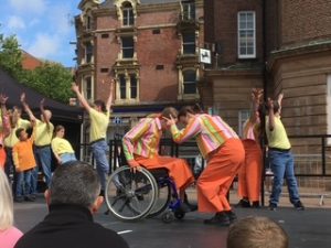 On the Piccadilly stage in Hanley town centre, the dancers are mid-performance, wearing different shades of bright yellow, orange and blue. Centre of the image, two dancers, one in her wheelchair, are bending forward, foreheads touching, with one cupping the other's head in her hands. Around them, the other dancers are holding both arms up in a straight V shape and looking up towards one of their hands. Behind them is the old Lloyd's Bank building.